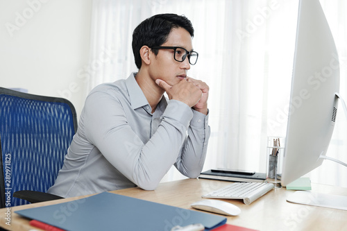 Pensive young Vietnamese entrepreneur reading document on screen of computer when working in his office