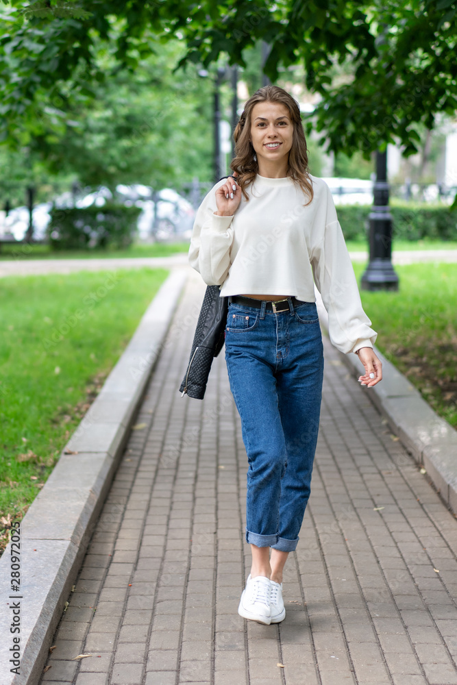 Beautiful European teen girl walking on pavement in city green park. Happy smile caucasian girl. Lifestyle, pullover, jeans, sneakers. It is interesting pose full length in fashionable look.