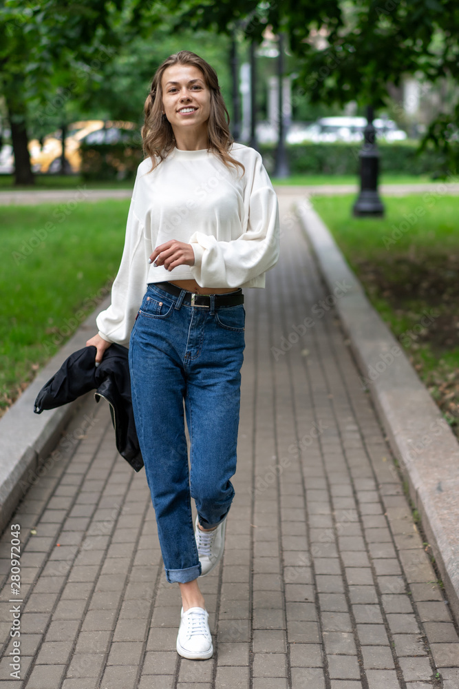 beautiful young European girl is having fun walking along sidewalk in city park. Happy caucasian girl. Lifestyle, pullover, jeans, sneakers. Waving arms, wide step, go fast.