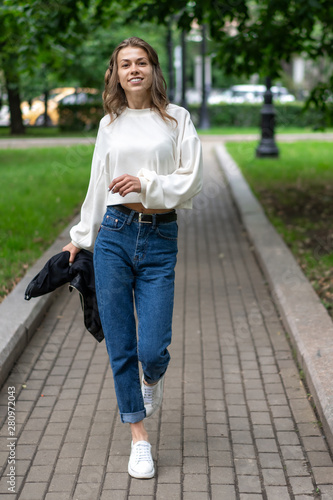 beautiful young European girl is having fun walking along sidewalk in city park. Happy caucasian girl. Lifestyle, pullover, jeans, sneakers. Waving arms, wide step, go fast. © Денис Бухлаев