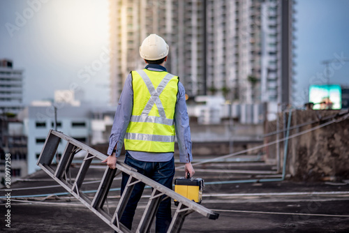 Αφίσα Asian maintenance worker man with safety helmet and green vest carrying aluminium step ladder and tool box at construction site
