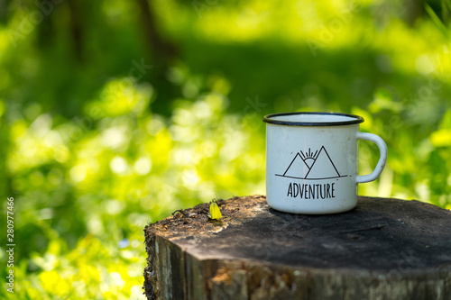 white enamel cup with mountains drawing and adventure text on aged stump in wild forest