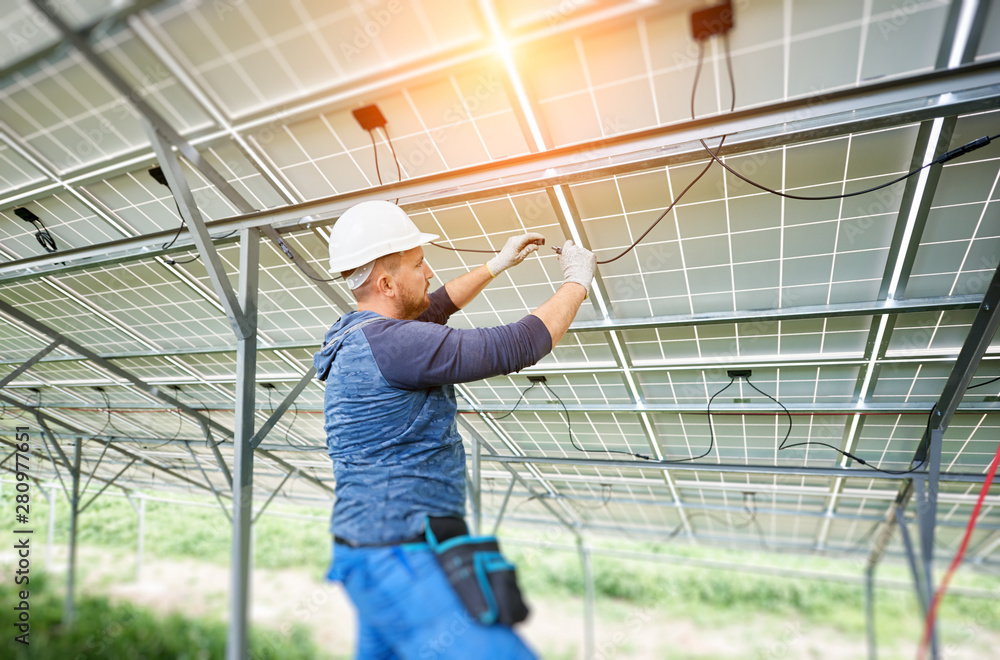 Young electrician connecting electrical cables inside the lit by sun solar modules. Installing and wiring of solar photo voltaic panel system. Alternative energy and profitable investment concept.