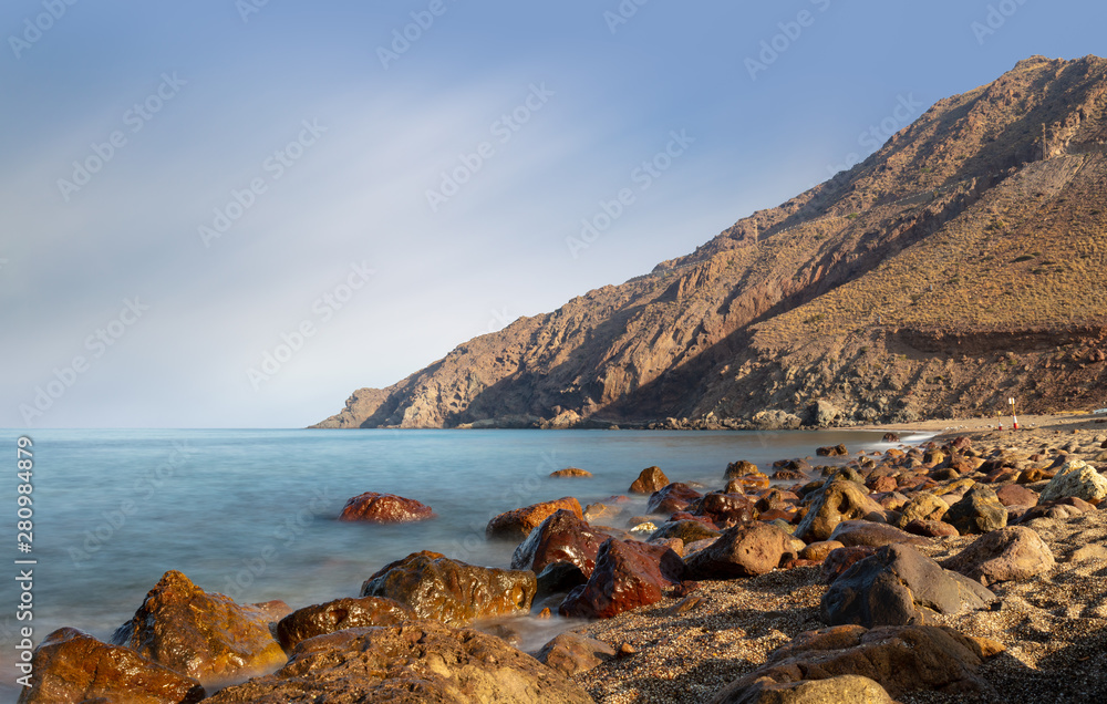 A long exposure on the Mediterranean with soft blue water and shiny stones on the beach. In the background the mountain at Cabo de Gata at Almeria in the morning light.
