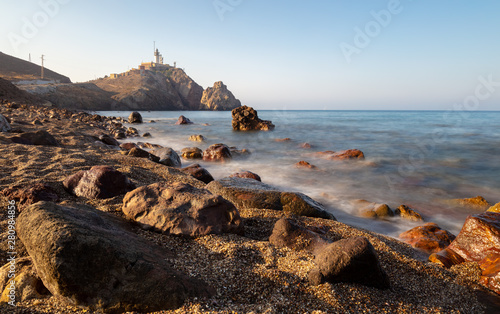 A long exposure on the Mediterranean with soft water and shiny stones on the beach. In the background the lighthouse Cabo de Gata at Almeria in the morning light.