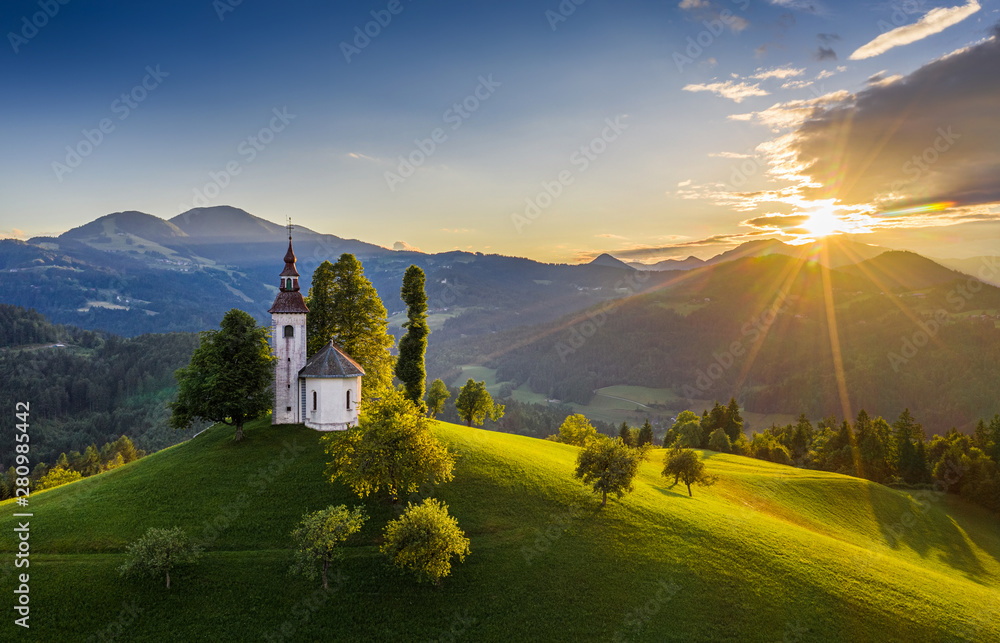Skofja Loka, Slovenia - Aerial view of the beautiful hilltop Sveti Tomaz (Saint Thomas) church with a warm summer sunset, clear blue sky and Julian Alps at the background - obrazy, fototapety, plakaty 