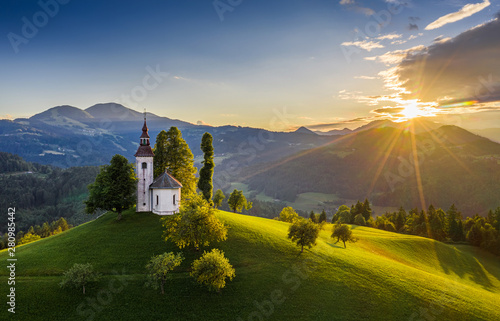 Skofja Loka, Slovenia - Aerial view of the beautiful hilltop Sveti Tomaz (Saint Thomas) church with a warm summer sunset, clear blue sky and Julian Alps at the background