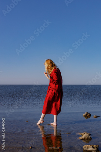 Blonde woman in a red long linen dress standing in the sea in the morning