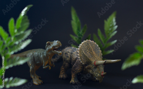 Realistic figures of the tyrannosaurus and triceratops dinosaurs under juicy green leaves on the black background