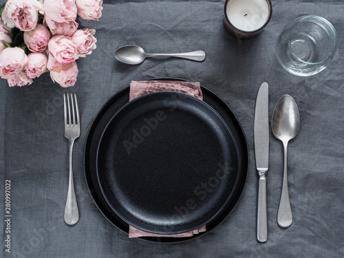 Beautiful table setting on gray linen tablecloth. Festive table setting for wedding dinner with pink spray roses and scented cabdle. Holiday dinner with black plates. Dinner, utensil. Copy space
