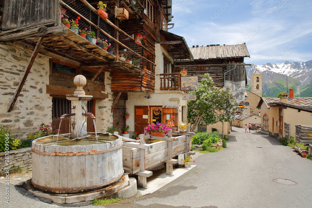Traditional wooden houses, a traditional fountain and the church in Saint Veran village, with mountain range covered with snow in the background, Queyras Regional Natural Park, Southern Alps, France
