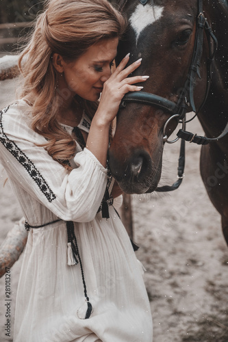 Beautiful and ladylike young woman wearing the dress is embracing and stroking the horse on the ranch. An attractive rider is posing outdoors near the saddle. Summertime, nature landscape, countryside