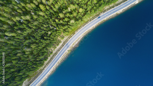 Beautiful aerial view of road between green summer forest and blue lake in Lapland. Car moving on road.