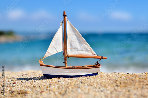 Photography Miniature toy sailboat on the beach against the background of the sea and the blue sky