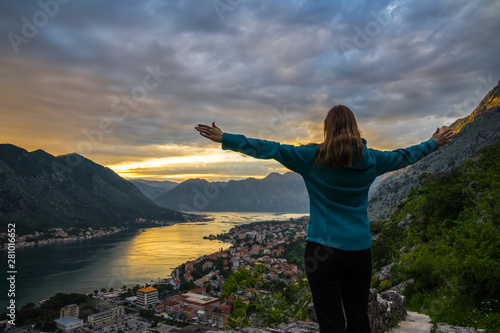 Montenegro, Beautiful young blonde woman standing with arms stretched wide above city of kotor bay in spectacular orange sunset twilight atmosphere in summer