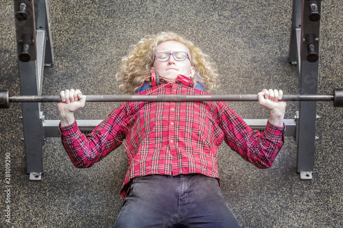 Curly blond in a plaid shirt strenuously raises the bar in the gym. photo