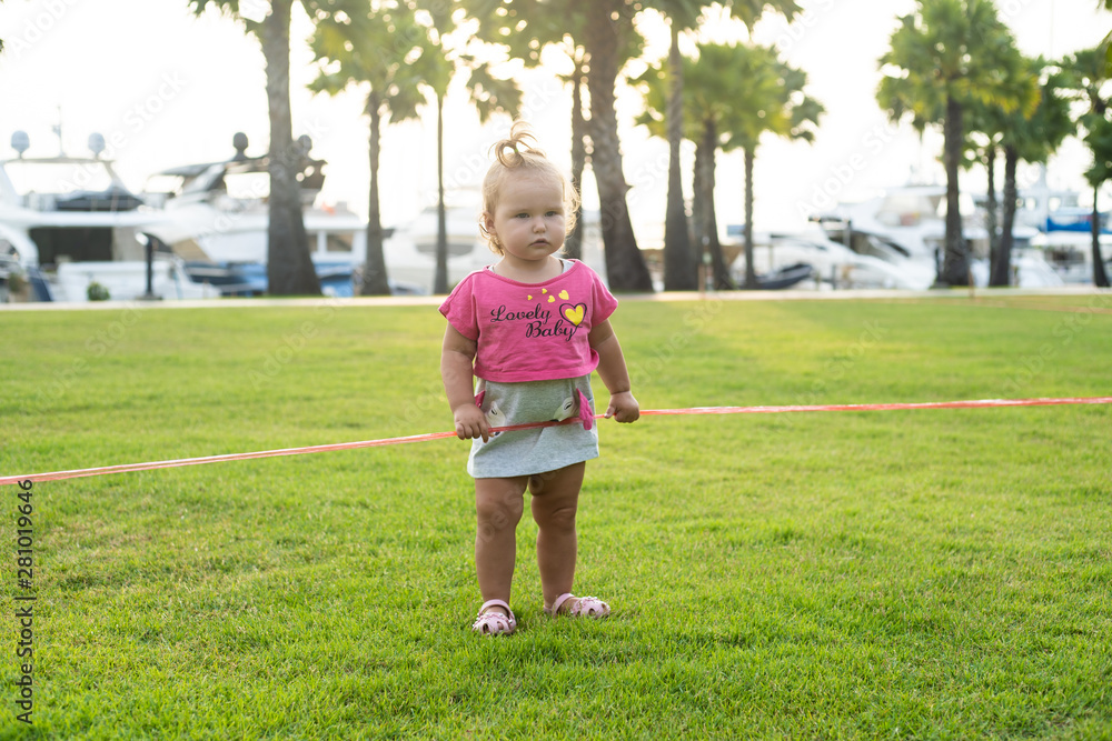 A little girl of 1 year old running on a green meadow in a park among palm trees and cutting a red ribbon