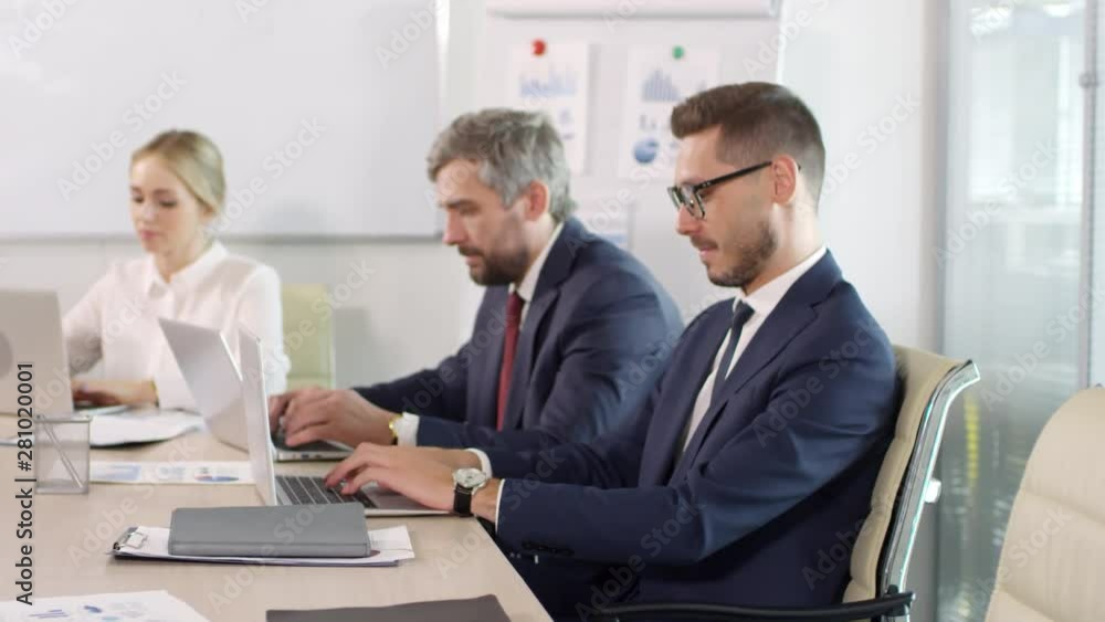 Dolly-in portrait shot of successful young businessman in suit and glasses working on laptop in conference room during meeting with male and female executives, then posing for camera and smiling Dolly-in portrait shot of successful young businessman in suit and glasses working on laptop in conference room during meeting with male and female executives, then posing for camera and smiling