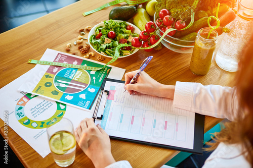 Woman dietitian in medical uniform with tape measure working on a diet plan sitting with different healthy food ingredients in the green office on background. Weight loss and right nutrition concept photo