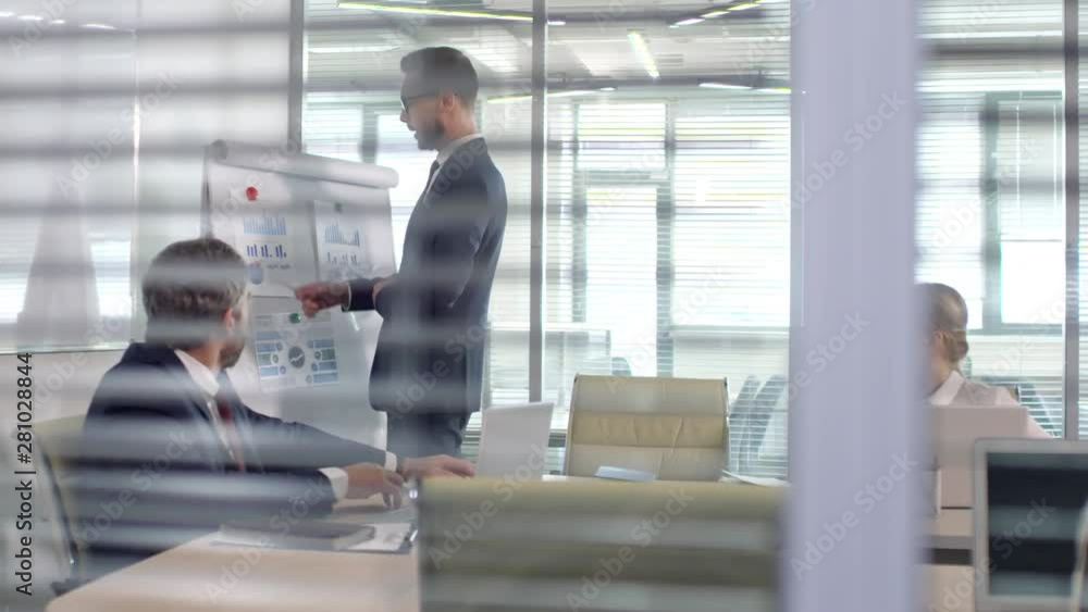 Tracking shot of businessman in suit and glasses delivering financial presentation to businesswoman and middle-aged male executive sitting in conference room Tracking shot of businessman in suit and glasses delivering financial presentation to businesswoman and middle-aged male executive sitting in conference room