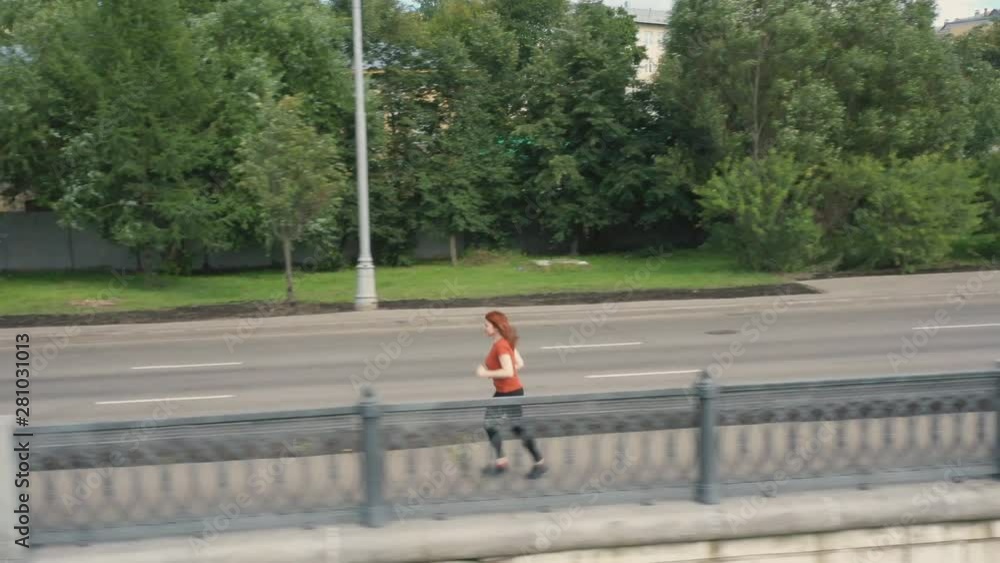 Shooting from the air red-haired sports woman enthusiastically running along the embankment of the city river Shooting from the air red-haired sports woman enthusiastically running along the embankment of the city river