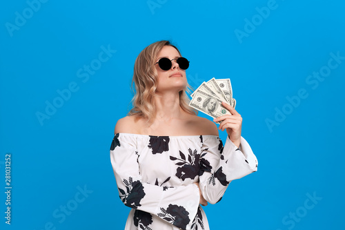Portrait of a girl with curly blond hair in a white dress and dark sunglasses standing on a blue background. Happy model fans herself with a bundle of dollars with dreamy look.