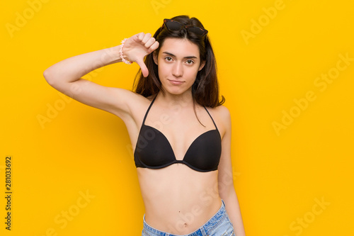 Young brunette woman wearing a bikini against yellow background showing a dislike gesture, thumbs down. Disagreement concept.