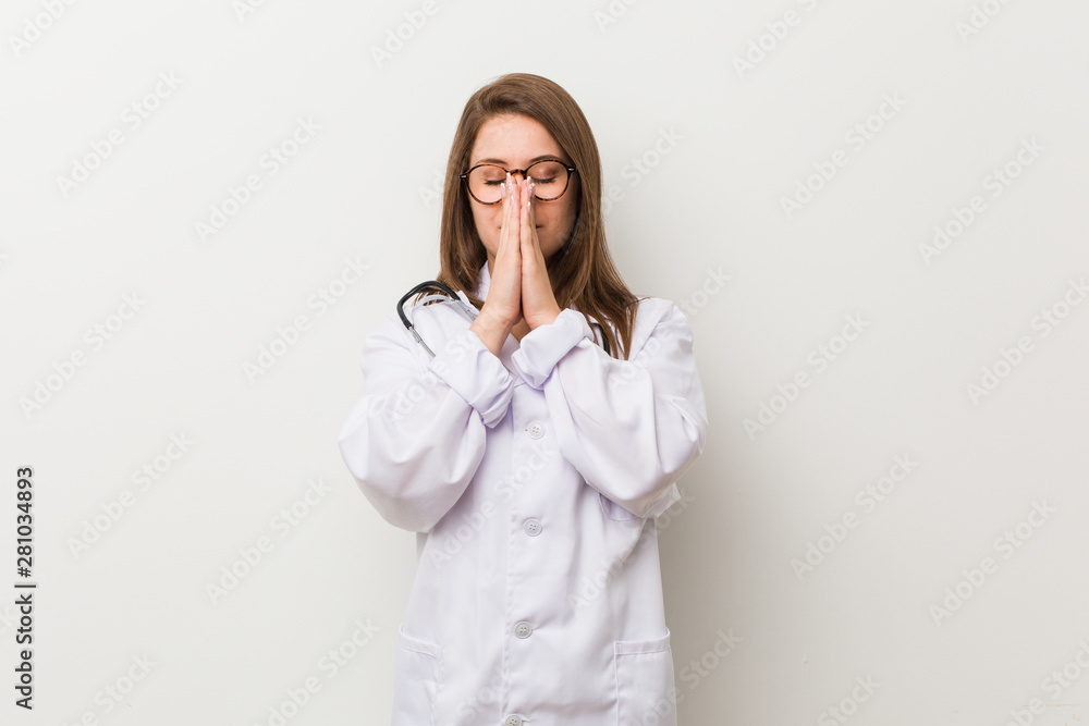 Young doctor woman against a white wall holding hands in pray near mouth, feels confident. Young doctor woman against a white wall holding hands in pray near mouth, feels confident.