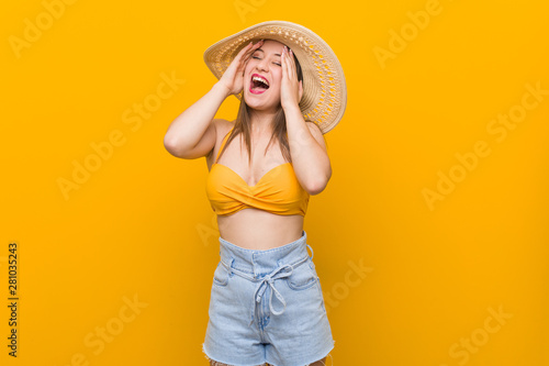 Young caucasian woman wearing a straw hat, summer look laughs joyfully keeping hands on head. Happiness concept.