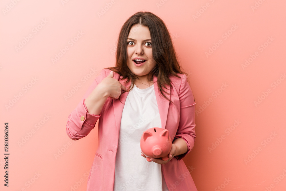 Young plus size curvy business woman holding a piggy bank surprised pointing at himself, smiling broadly. Young plus size curvy business woman holding a piggy bank surprised pointing at himself, smiling broadly.