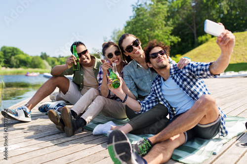 leisure, picnic and technology concept - friends with drinks taking selfie by smartphone on lake pier in summer park © Syda Productions