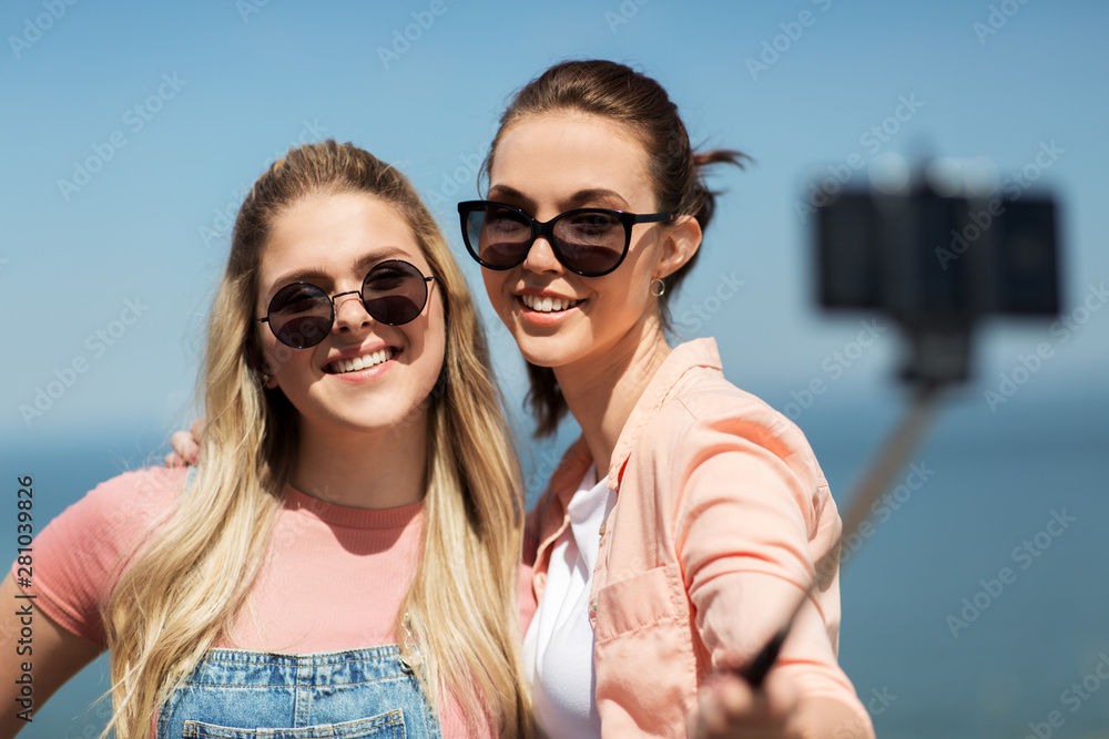 leisure and friendship concept - happy smiling teenage girls or best friends in sunglasses hugging and taking picture by smartphone on selfie stick at seaside in summer