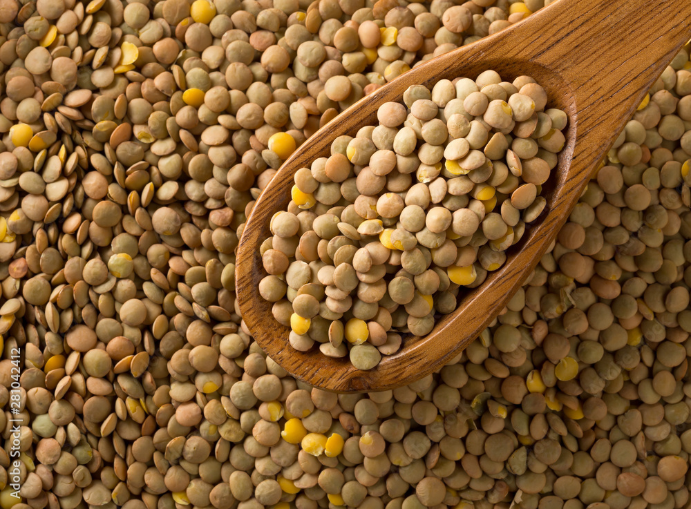 Raw, dry, uncooked brown lentil legumes in wooden spoon on lentil background flat lay top view from above Raw, dry, uncooked brown lentil legumes in wooden spoon on lentil background flat lay top view from above