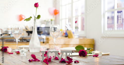 beautiful romantic set up dinner table at home with flowers and rose petals surrounding love letter on wooden desk on valentine day. kitchen in background. candles warm up the room with light up fire