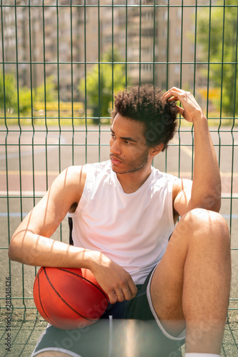 Young serious restful basketball player with ball sitting by fence of playground © pressmaster