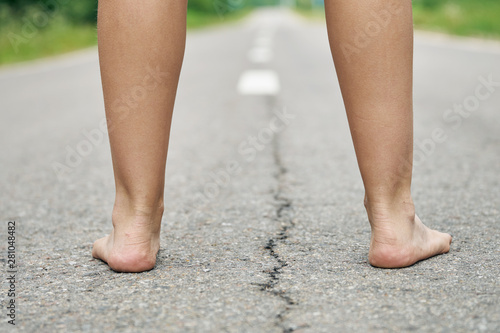 Rear view of bare feet of a young girl standing on the asphalt road close up. Dividing road lines are visible far away between legs.