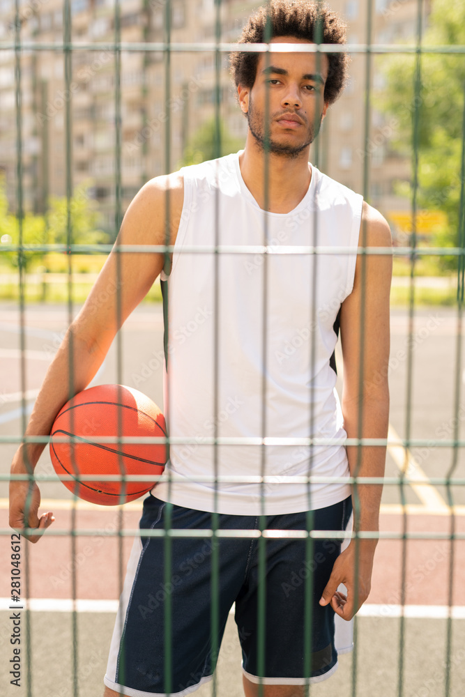 Young serious active player with ball looking at you through fence Young serious active player with ball looking at you through fence