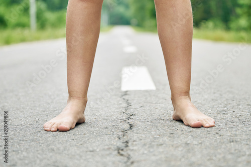 Front view of bare feet of a young girl walking along the asphalt road close up. Dividing road lines are visible far away between legs.