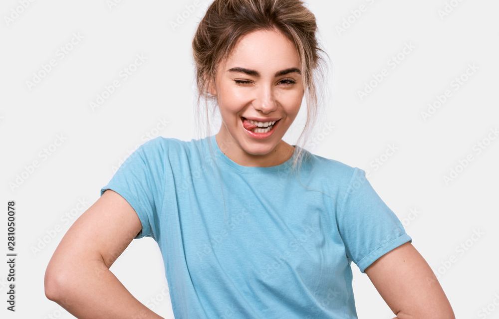 Positive Caucasian young woman smiling broadly, blinking with an eye, showing peace gesture and shows tongue while looking to the camera, standing against white studio background. Positive Caucasian young woman smiling broadly, blinking with an eye, showing peace gesture and shows tongue while looking to the camera, standing against white studio background.