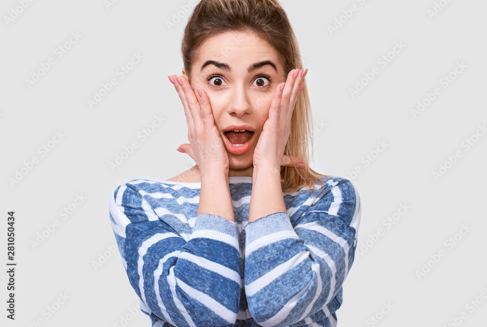 Closeup portrait of shocked young woman shouts holds cheeks by hands. Amazed female hears something pleasant, keeps hands on cheeks, isolated over white background with copy space for your text. Closeup portrait of shocked young woman shouts holds cheeks by hands. Amazed female hears something pleasant, keeps hands on cheeks, isolated over white background with copy space for your text.