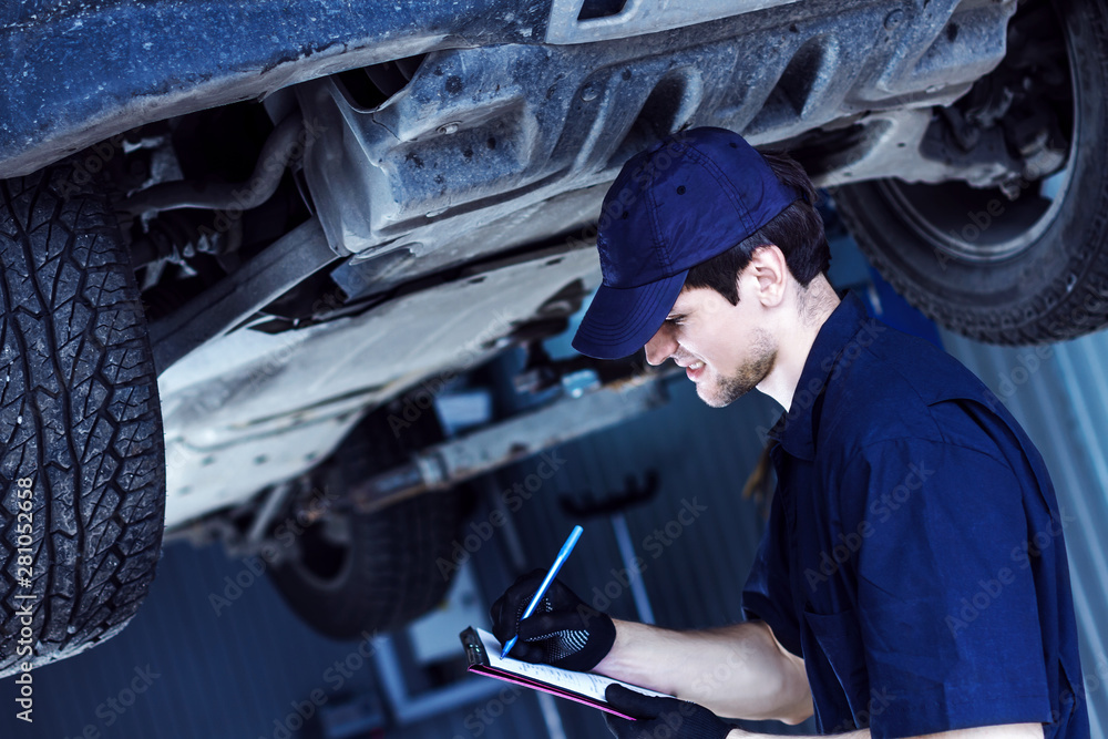 Mechanic in blue jumpsuit is repairing car at service station garage. Smiling repairman is holding job sheet for repairs of vehicle in workshop. Silver automobile on hydraulic lift is above. Mechanic in blue jumpsuit is repairing car at service station garage. Smiling repairman is holding job sheet for repairs of vehicle in workshop. Silver automobile on hydraulic lift is above.