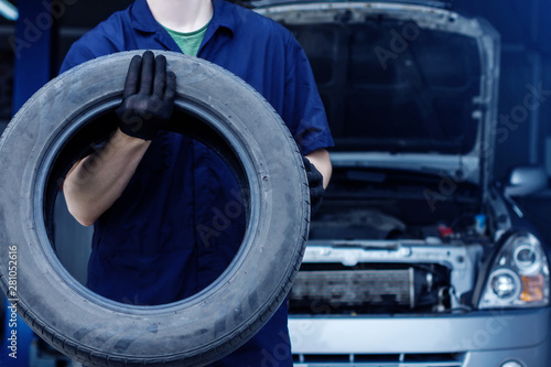 Mechanic in blue jumpsuit is repairing car at service station. Closeup of repairman hands are holding tyre from wheel in workshop. Vehicle with open hood on background. Tire fitting concept.