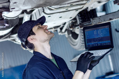 Mechanic is repairing car at service station. Repairman is conducting diagnostics and detecting problems by special software on laptop at workshop auto repair shop. Vehicle on hydraulic lift is above.