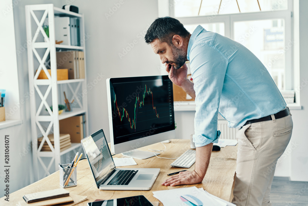 Middle age man in smart casual wear analyzing stock market data and talking on the phone while working in the office Middle age man in smart casual wear analyzing stock market data and talking on the phone while working in the office