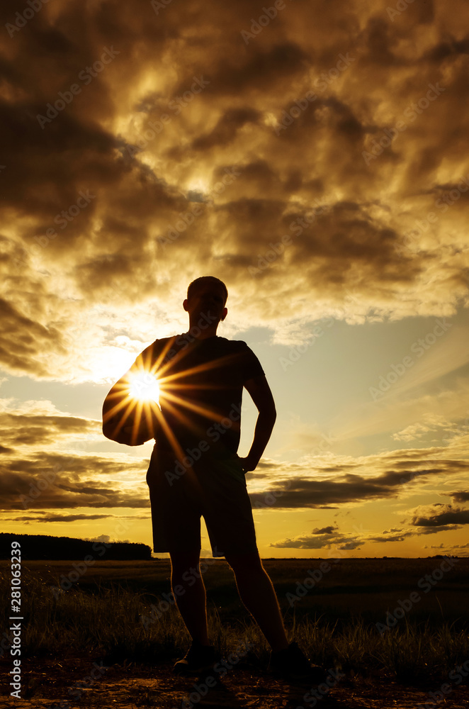 Silhouette of a man with a short haircut in a T-shirt and shorts with a ball. The concept of games with a ball in the fresh air, volleyball. Photo in warm sunset colors. Place for text. 