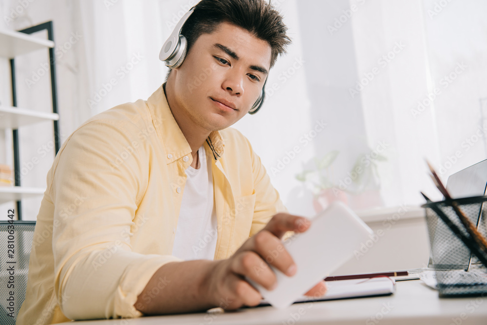 serious asian man in headphone looking at smartphone while sitting at table serious asian man in headphone looking at smartphone while sitting at table