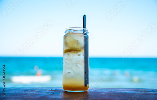 A jar of lemonade, in which float lemon and ice, with two black tubes, on the bar against the sea.