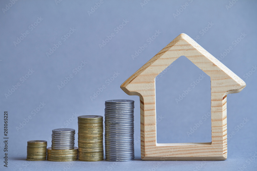 A wooden figurine of house with several columns of coins nearby on gray background, the concept of buying or renting a building, selective focus A wooden figurine of house with several columns of coins nearby on gray background, the concept of buying or renting a building, selective focus