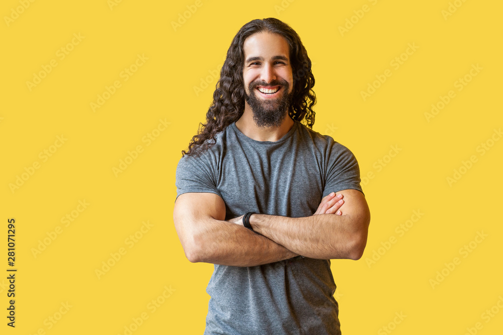 Portrait of handsome happy bearded young man with long curly hair in grey tshirt standing with crossed arms and looking at camera with toothy smile. indoor studio shot isolated on yellow background. Portrait of handsome happy bearded young man with long curly hair in grey tshirt standing with crossed arms and looking at camera with toothy smile. indoor studio shot isolated on yellow background.