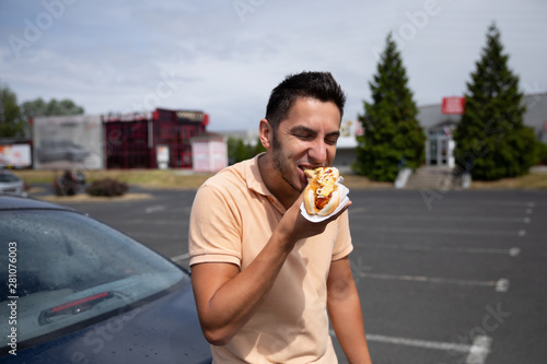 Fotografie Handsome young brunette man eating hot dog in the parking lot near the gas station